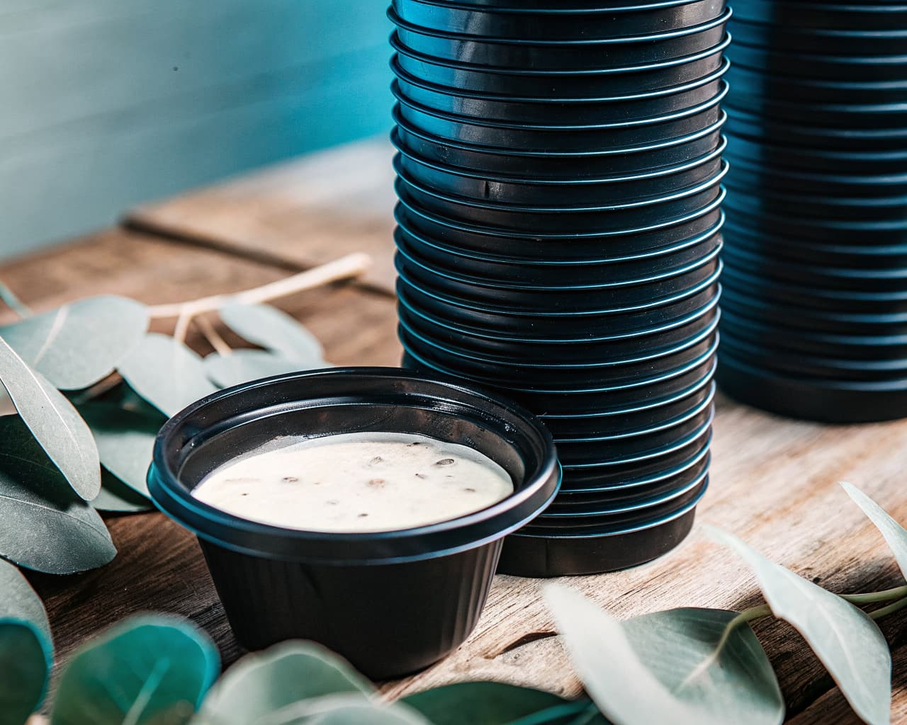 Stacked PHA biodegradable 2 oz black souffle cups with a tight-fitting lid and dressing inside, on a wooden surface with eucalyptus leaves — compostable drop-in replacement for conventional plastic portion cups