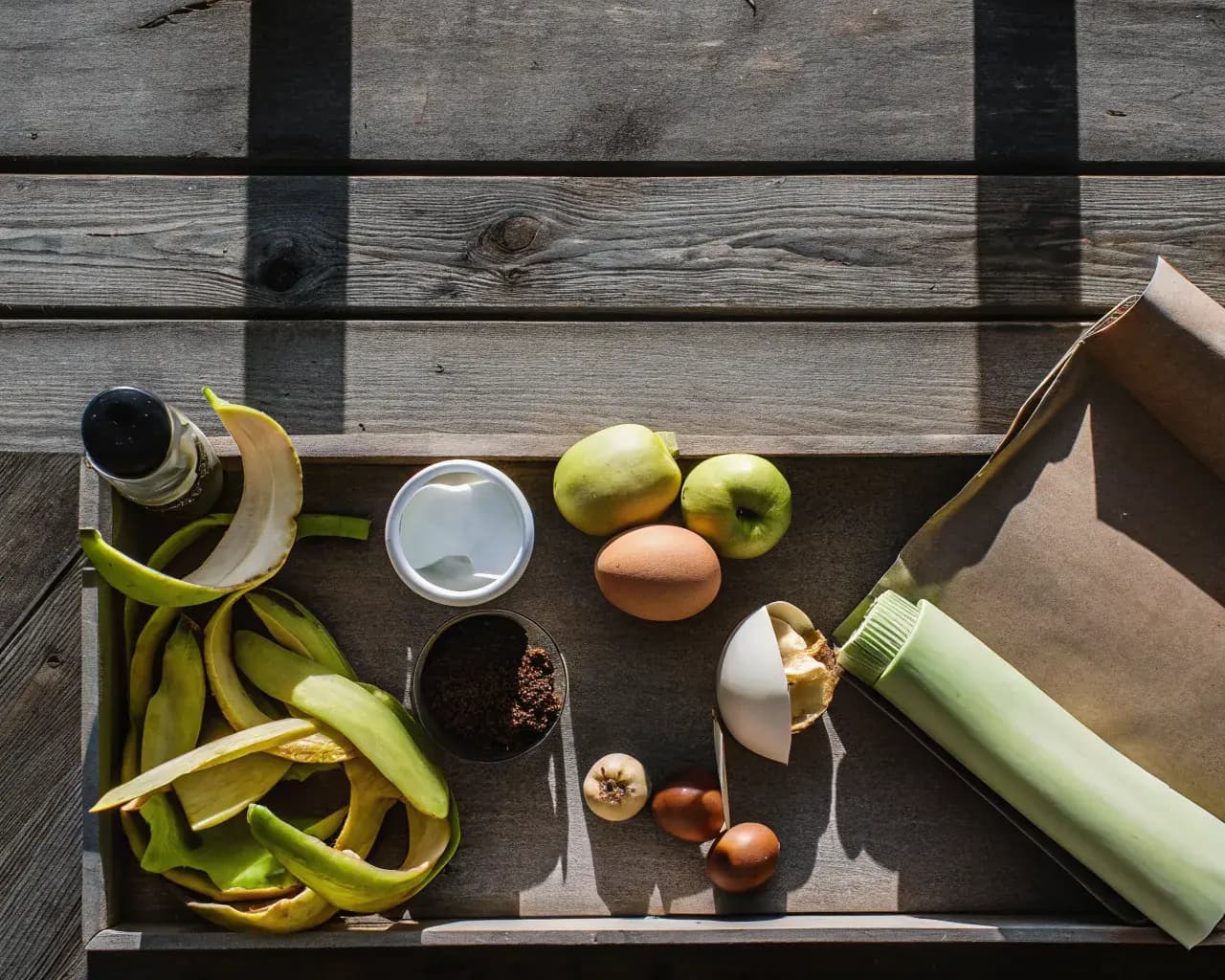Flat lay of compostable kitchen scraps including banana peels, apple cores, coffee grounds, eggshells, and cardboard arranged on a wooden surface